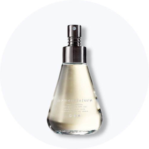 Her ses et billede af en parfumeflakon fra Nomenclature placeret centralt mod en lys, rund baggrund. Flakonen er i klart glas med sølvfarvet spraytop og har teksten “nomenclature” på fronten.