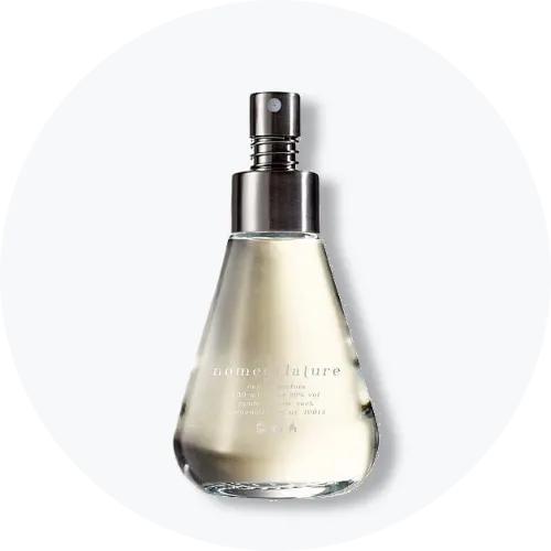 Her ses et billede af en parfumeflakon fra Nomenclature placeret centralt mod en lys, rund baggrund. Flakonen er i klart glas med sølvfarvet spraytop og har teksten “nomenclature” på fronten.
