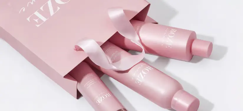 Her ses tre ROZE Avenue produkter, som kommer ud af en lyserød pose. Varene og posen ligger på et hvidt bord.