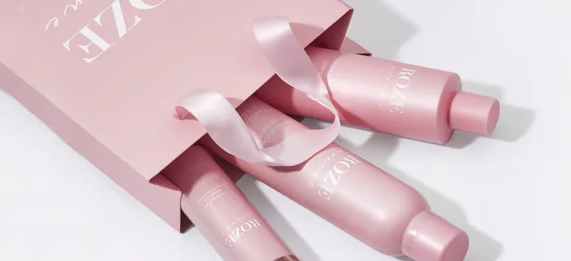 Her ses tre ROZE Avenue produkter, som kommer ud af en lyserød pose. Varene og posen ligger på et hvidt bord.