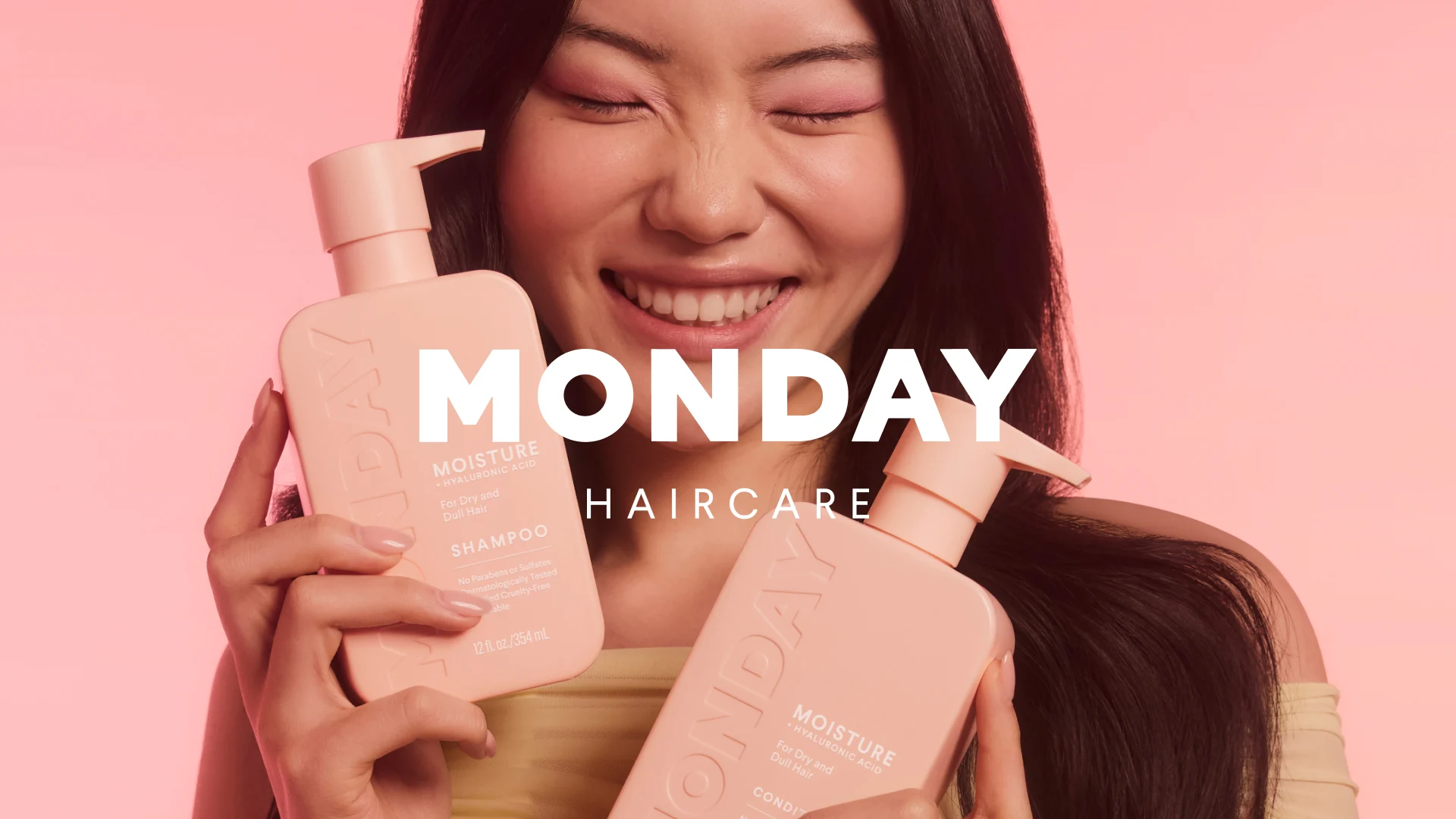 Her ses et kampagnebillede for MONDAY Haircare med en smilende person med langt mørkt hår, der holder MONDAY Moisture Shampoo og Conditioner i rosa pumpeflasker. På billedet står teksten “MONDAY Haircare”.