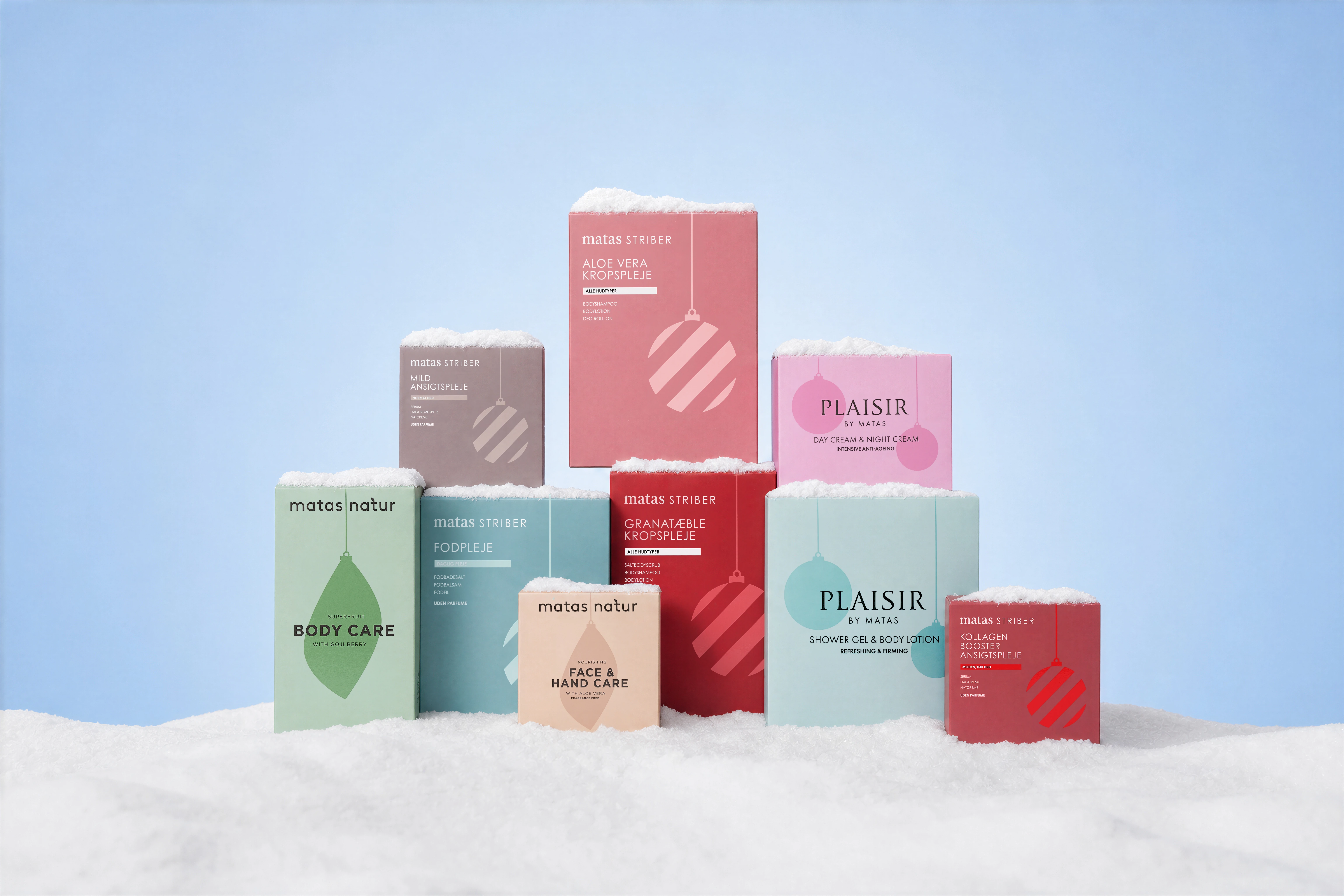 Et festligt Matas og Plaisir produktdisplay i sne, med kropspleje, ansigtspleje og fodplejesæt, dekoreret med julekugler.