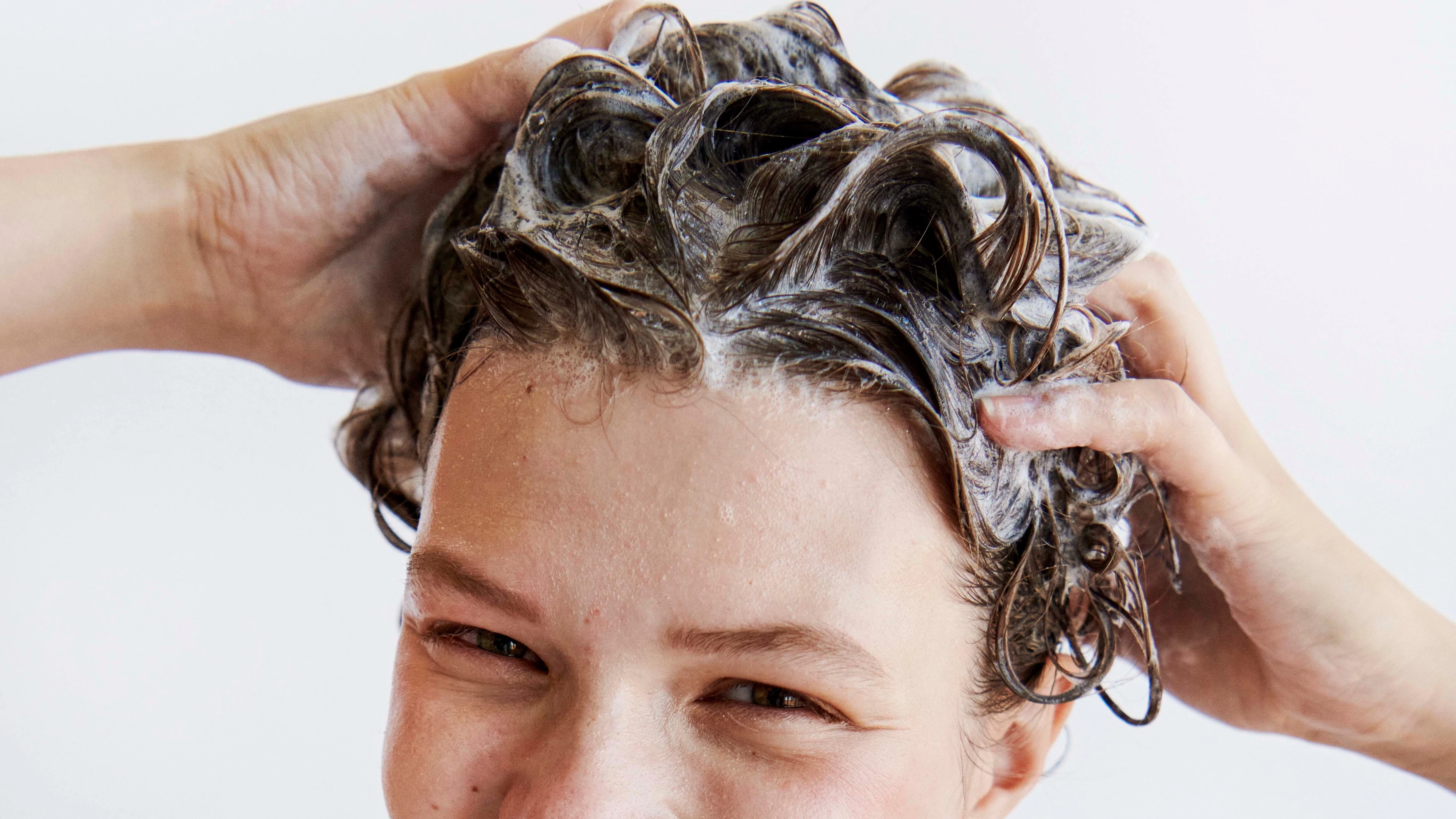 Smilende person vasker vådt, krøllet hår med shampoo og er pakket ind i et lilla håndklæde.