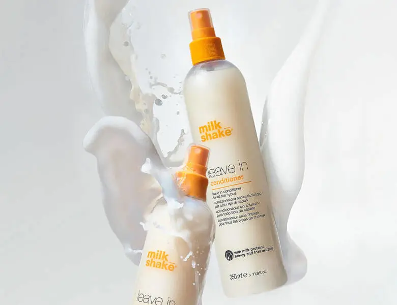 Her ses et billede med to flasker fra milk_shake placeret mod en lys baggrund med væske, der sprøjter omkring dem. På flaskerne står teksten “milk_shake leave in conditioner”. Flaskerne er i lyse nuancer med orange sprayhoveder.