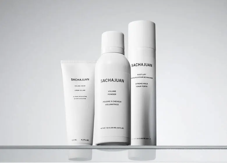 Her ses et billede med tre hårstylingprodukter fra SACHAJUAN placeret på en lys, reflekterende overflade mod en lys baggrund. På produkterne står teksten “SACHAJUAN”, “Volume Cream”, “Volume Powder” og “Root Lift”. Produkterne er i hvide emballager.
