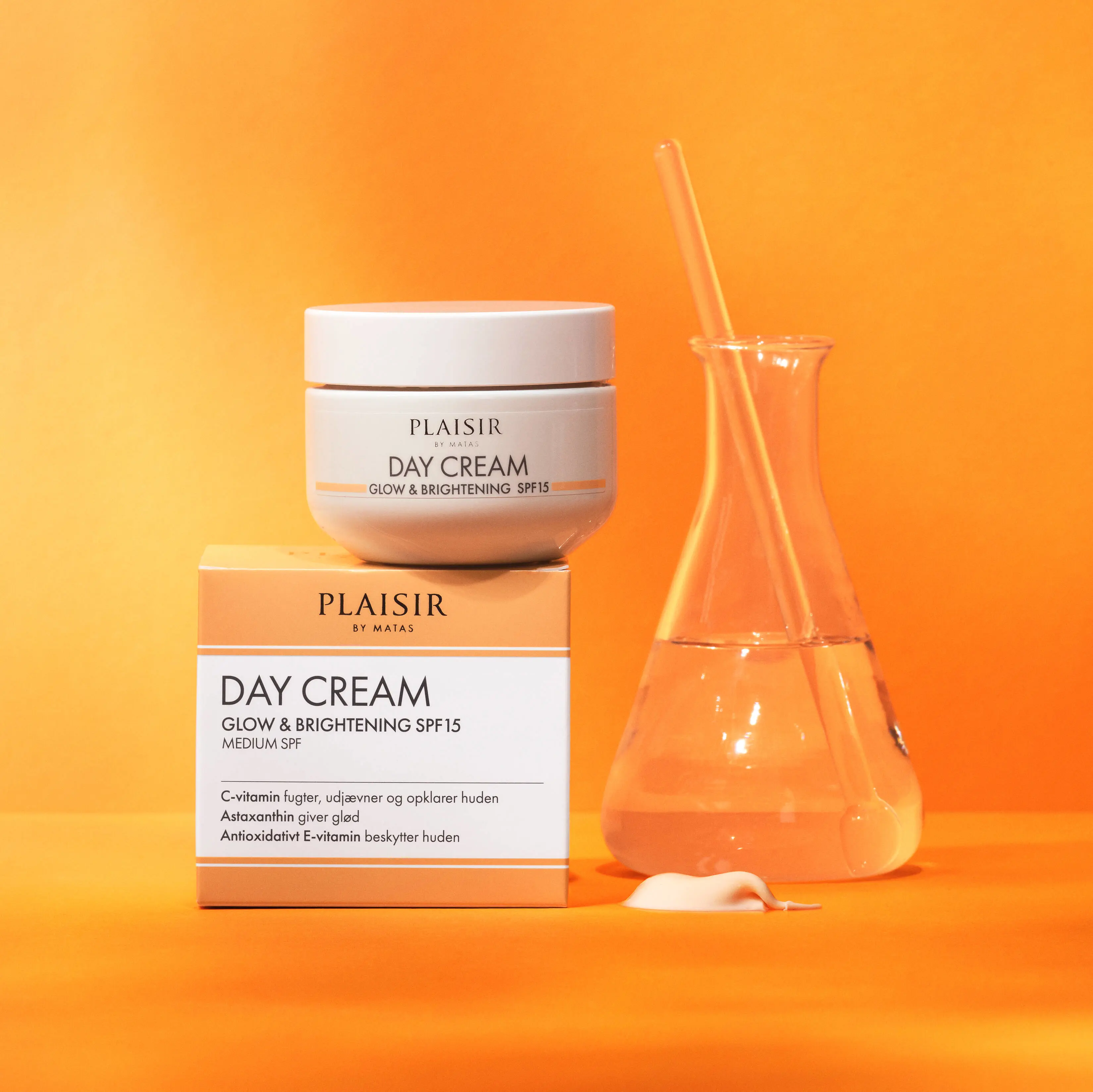 Produktbillede af “Plaisir by Matas Day Cream Glow & Brightening SPF15” på en varm orange baggrund. En hvid dagcreme-krukke står oven på den tilhørende produktæske i hvide og lyse orange farver. Ved siden af produkterne står en glasbeholder med klar væske og en glasstav. Foran ligger en lille klat creme. Billedet har et rent og luksuriøst udtryk med fokus på fugtgivende og glødgivende hudpleje.