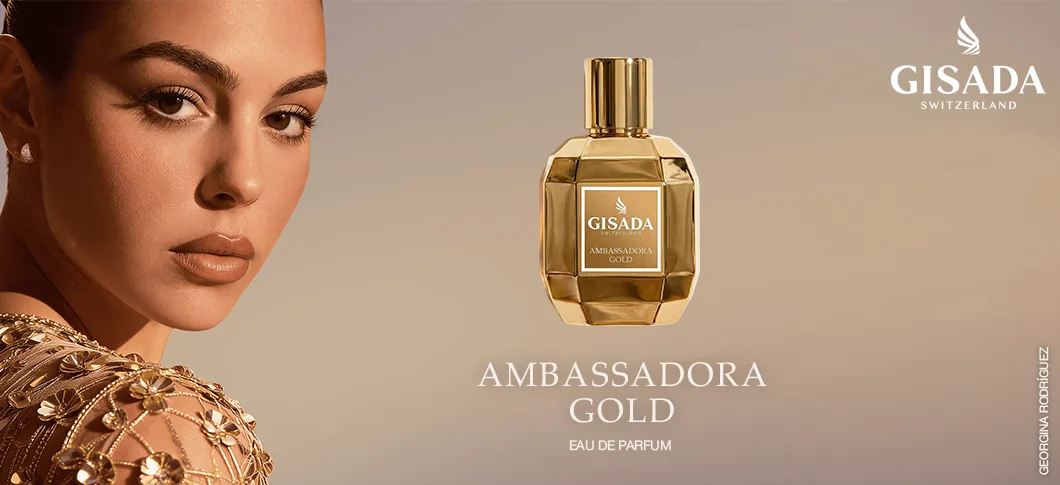 Her ses et billede af en person mod en varm beige baggrund ved siden af en parfumeflakon fra Gisada. På billedet står teksterne “GISADA SWITZERLAND” og “AMBASSADORA GOLD EAU DE PARFUM”. Personen er iført guldfarvede smykker, og parfumeflakonen har guldfarvede detaljer.