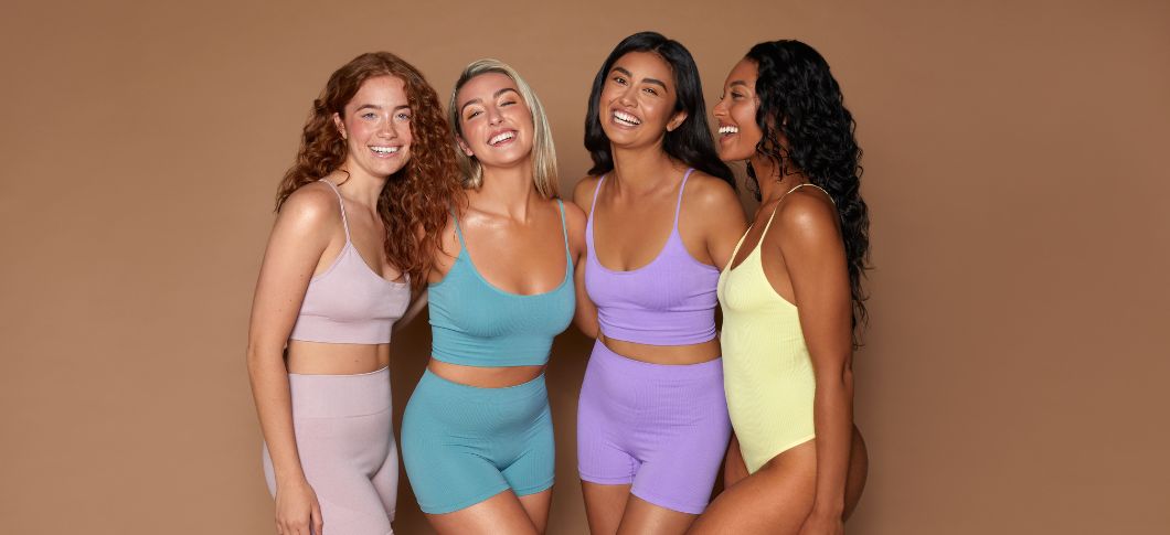 4 kvinder står i træningstøj på en brun baggrund. De ser glade ud og har forskellige farver på tøjet. 