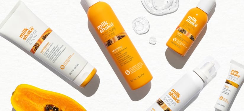 Her ses et billede med flere hårplejeprodukter fra milk_shake placeret spredt på en lys overflade. På produkterne står blandt andet “milk_shake moisture plus shampoo” og “conditioner”. Produkterne er primært i orange og hvide emballager, og der ses frugt og vanddråber omkring dem.