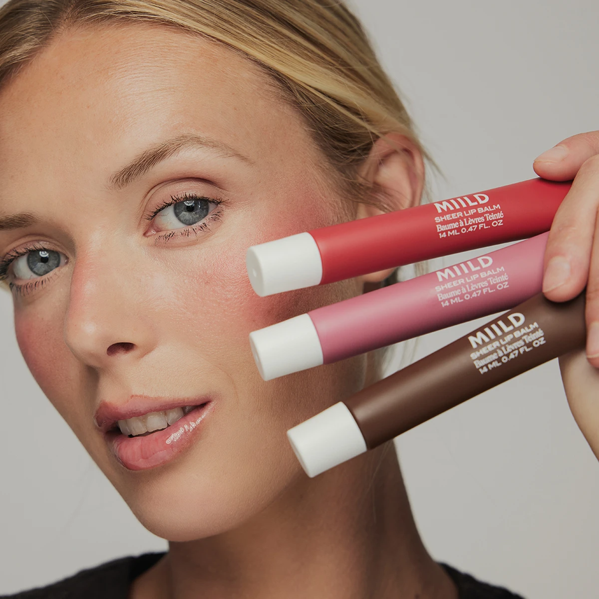 Model holder tre MIILD Sheer Lip Balm, Baume à Lèvres Teinté, 14 ML 0.47 FL. OZ, i rød, pink og brun, med rødmede kinder og glinsende læber.