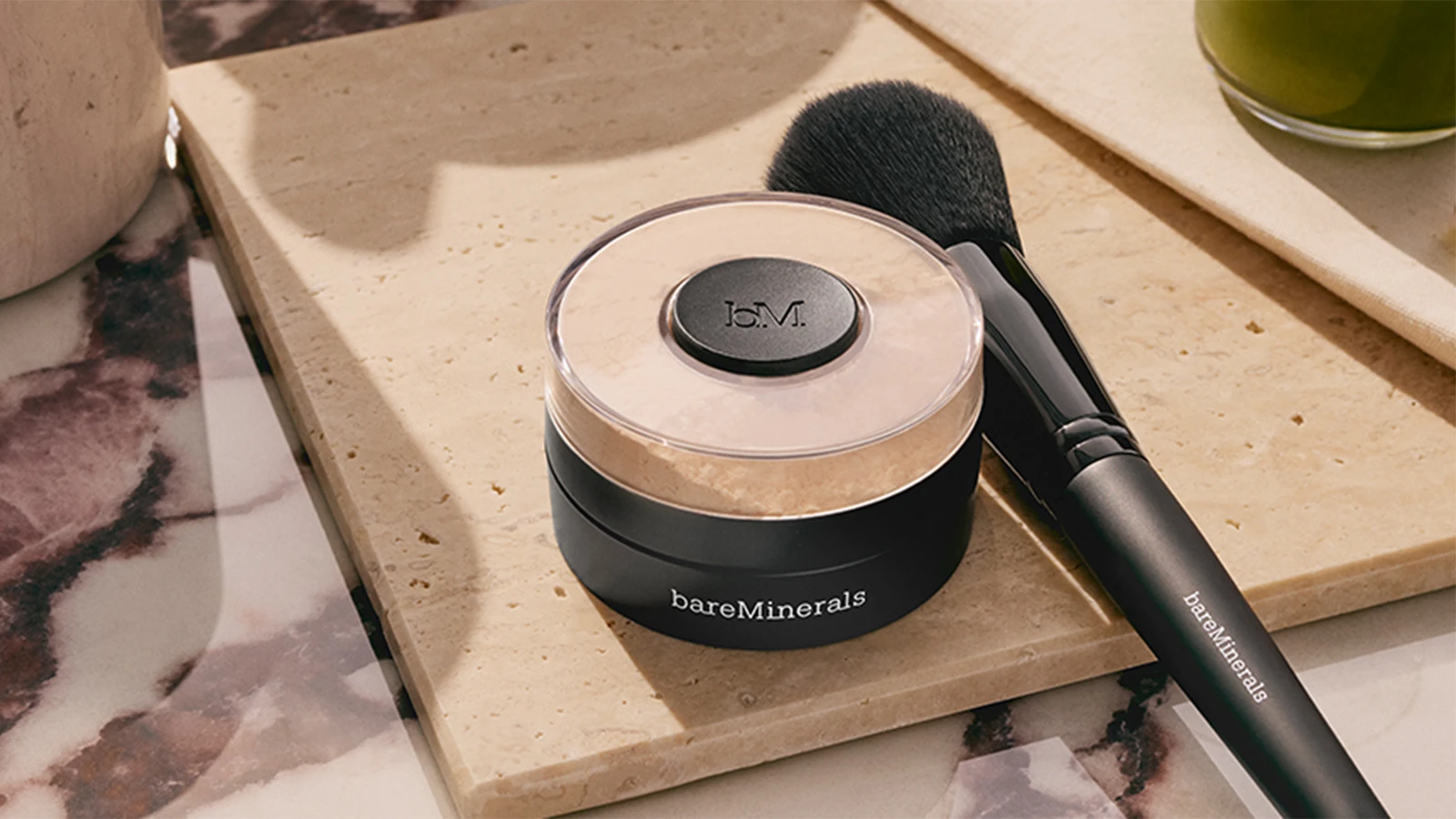 bareMinerals løst pudder med 'JCM.' logo og matchende makeupbørste, på beige stenflade.