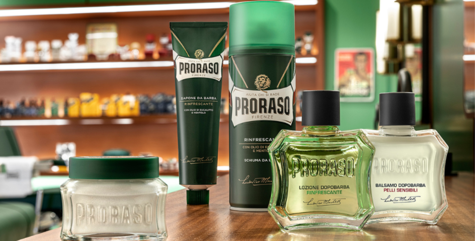 Proraso barberingsprodukter står opstillet på et træbord i en barbershop, herunder barbersæbe, barberskum, aftershave og balm i grøn og hvid emballage.