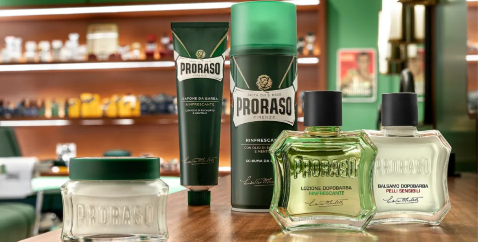 Proraso barberingsprodukter står opstillet på et træbord i en barbershop, herunder barbersæbe, barberskum, aftershave og balm i grøn og hvid emballage.