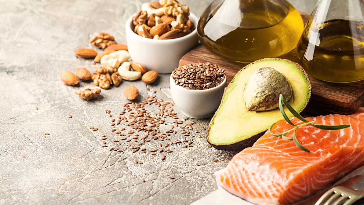 Her ses avocado, laks, nødder, frø og olie arrangeret på et bord.