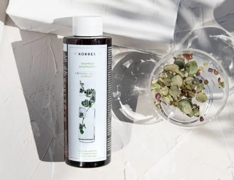 Her ses en hvid baggrund med et liggende produkt til håret. Billedet er taget oppefra og der er et glas med nogle grønne planter. 