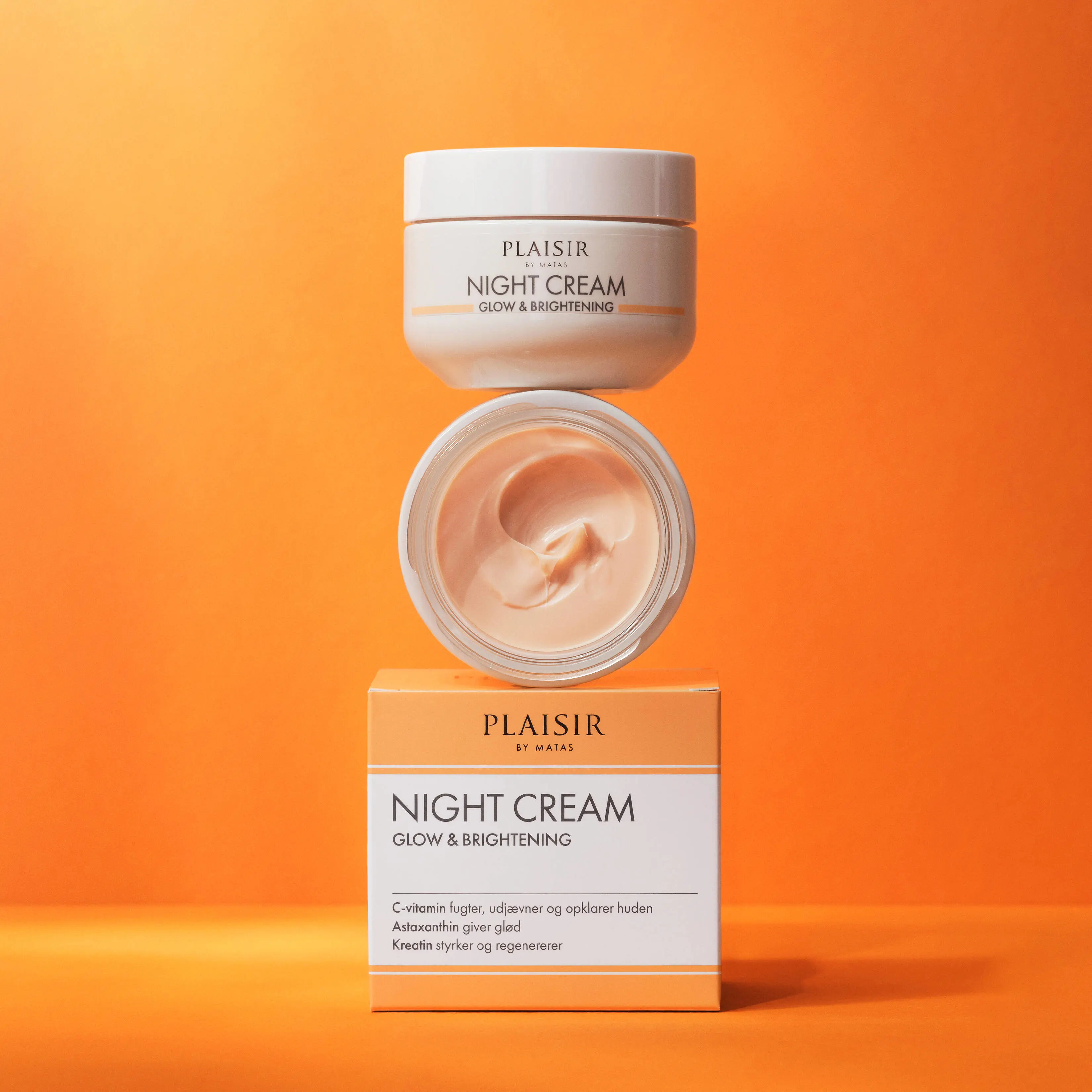 Produktbillede af “Plaisir by Matas Night Cream Glow & Brightening” på en varm orange baggrund. Nederst står den tilhørende produktæske i hvide og lyse orange farver. Ovenpå æsken står en åben krukke med synlig creme i et blødt swirl-mønster, og over den ses låget med produktnavnet. Billedet har et rent og luksuriøst udtryk med fokus på plejende natcreme og glødgivende hudpleje.