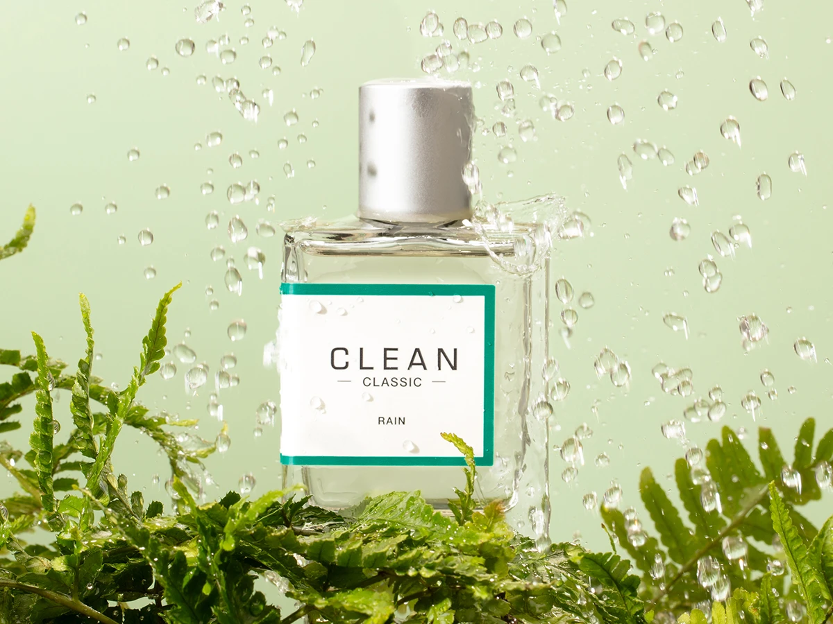 Her ses CLEAN Classic Rain i en transparent parfumeflakon med sølvfarvet låg. Flakonen er omgivet af grønne planter og vanddråber mod en lysegrøn baggrund.
