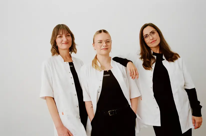 Tre kvinder i hvide kitler og sorte trøjer med logoet "natas" står smilende foran en hvid baggrund.