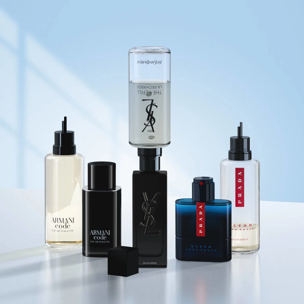 Fem luksusparfumer står samlet på en lys overflade foran en blød lyseblå baggrund. Produkterne fra Armani, Yves Saint Laurent og Prada vises sammen med flere refillflasker monteret til genopfyldning. I midten ses en sort YSL MYSLF-flaske med refill ovenpå, omgivet af Armani Code- og Prada Luna Rossa Ocean-dufte. Billedet har et moderne og eksklusivt udtryk med fokus på refillbare parfumer.