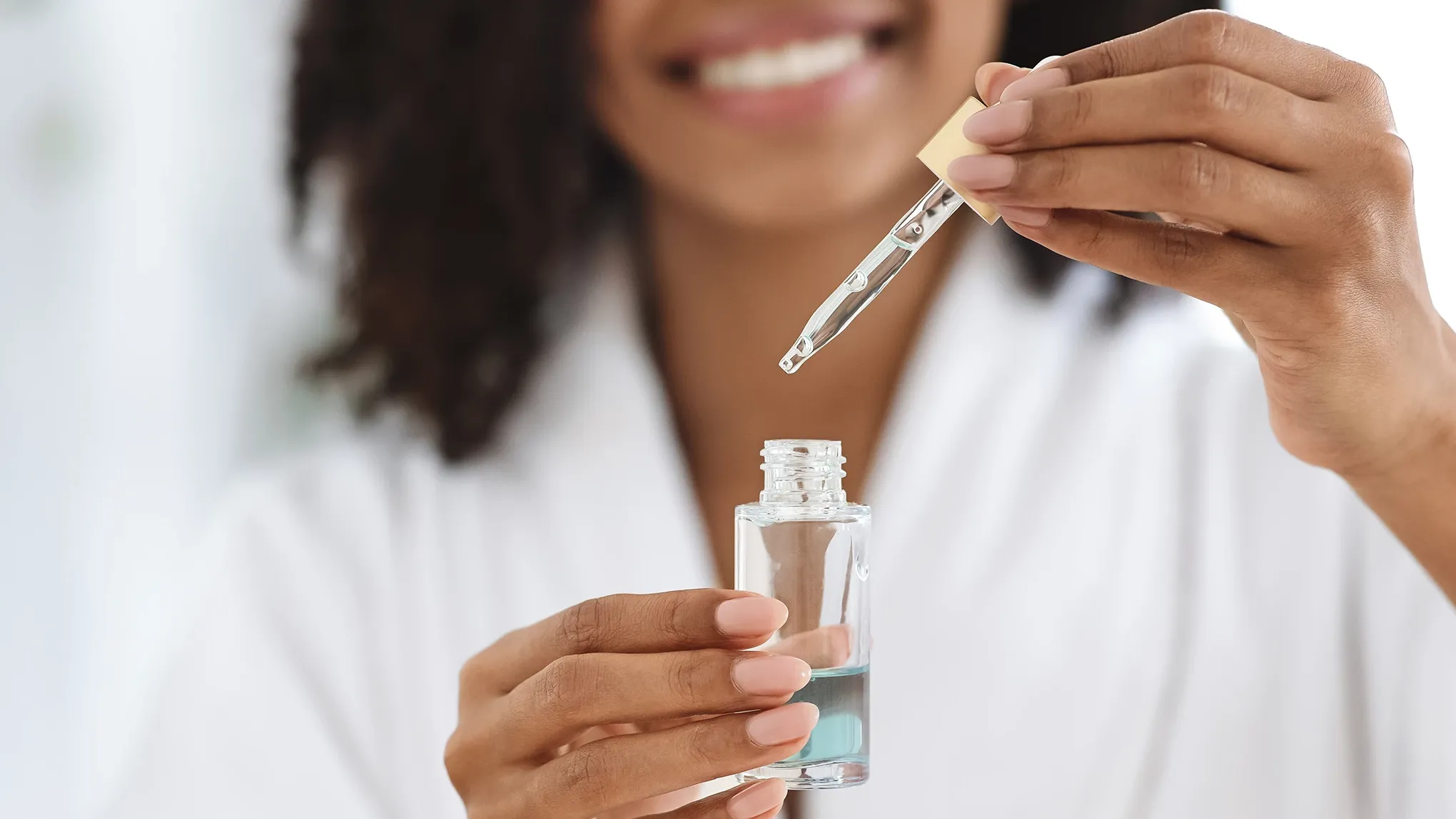 En suddig kvinna med leende och mörkt hår syns i bakgrunden bakom ett blått serum i en transparent, öppnad glasflaska.