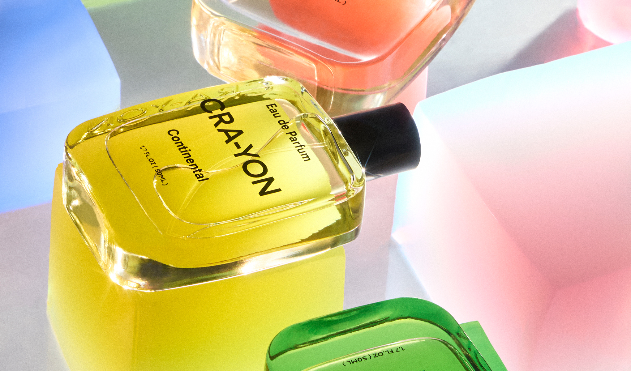 Useita CRA-YON-merkin hajuvesipulloja. Pääasiallinen pullo on keltainen ja siinä on teksti ”CRA-YON Eau de Parfum Continental”. Taustalla on värikkäitä, läpinäkyviä kuutioita, jotka luovat kirkkaan ja leikkisän tunnelman.