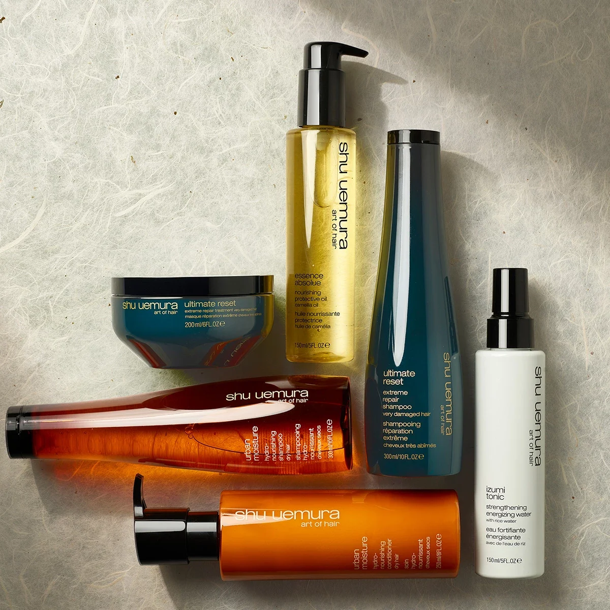 En samling hårvårdsprodukter från Shu Uemura, inklusive schampo, balsam, hårmask, hårolja och stärkande tonic. Produkterna har färgglada flaskor och burkar i orange, blått, vitt och guld.