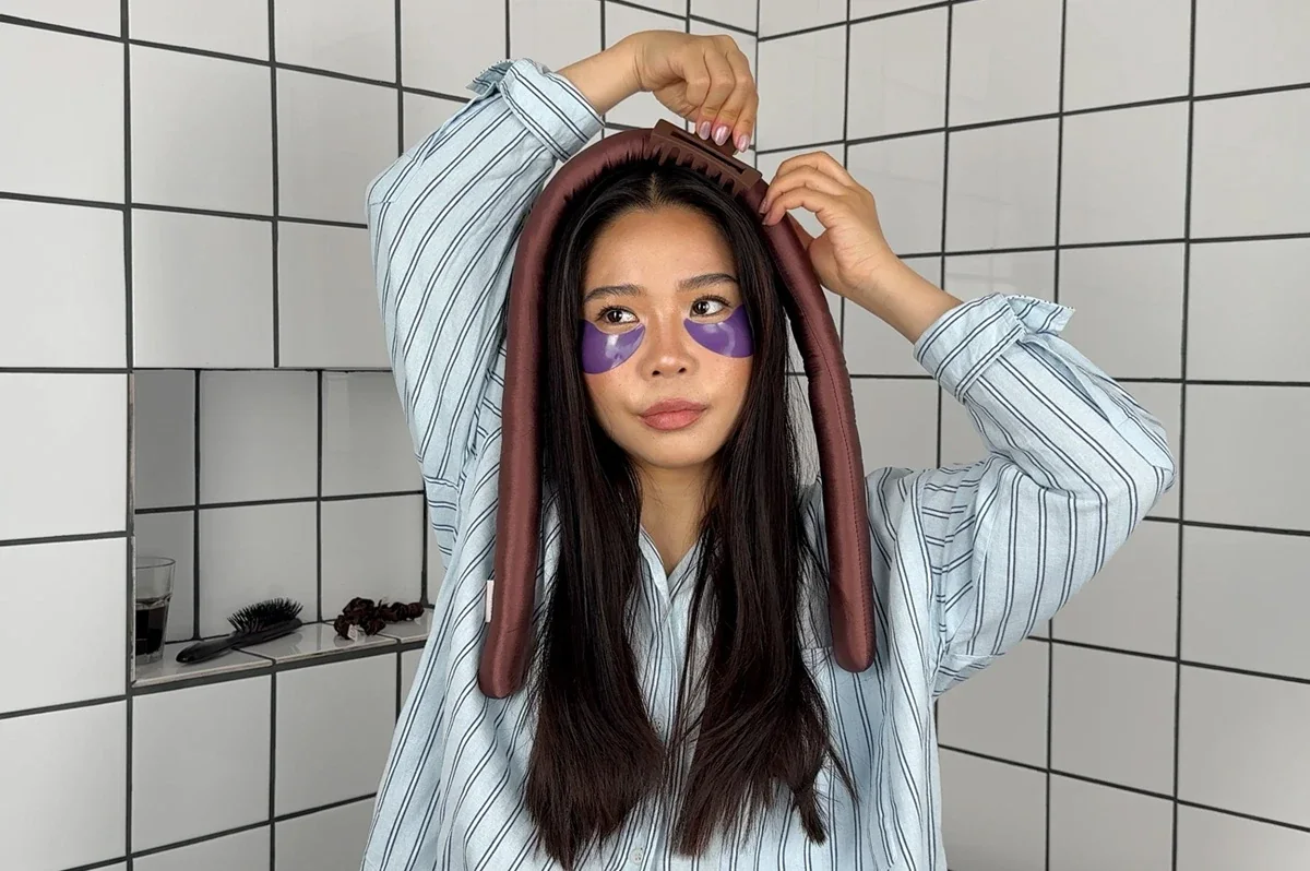 Person fester et brunt varmefritt krøllebånd på hodet med en hårklemme før varmefri hårstyling. Personen har langt, mørkt hår, lilla øyemasker og en lyseblå stripete skjorte.