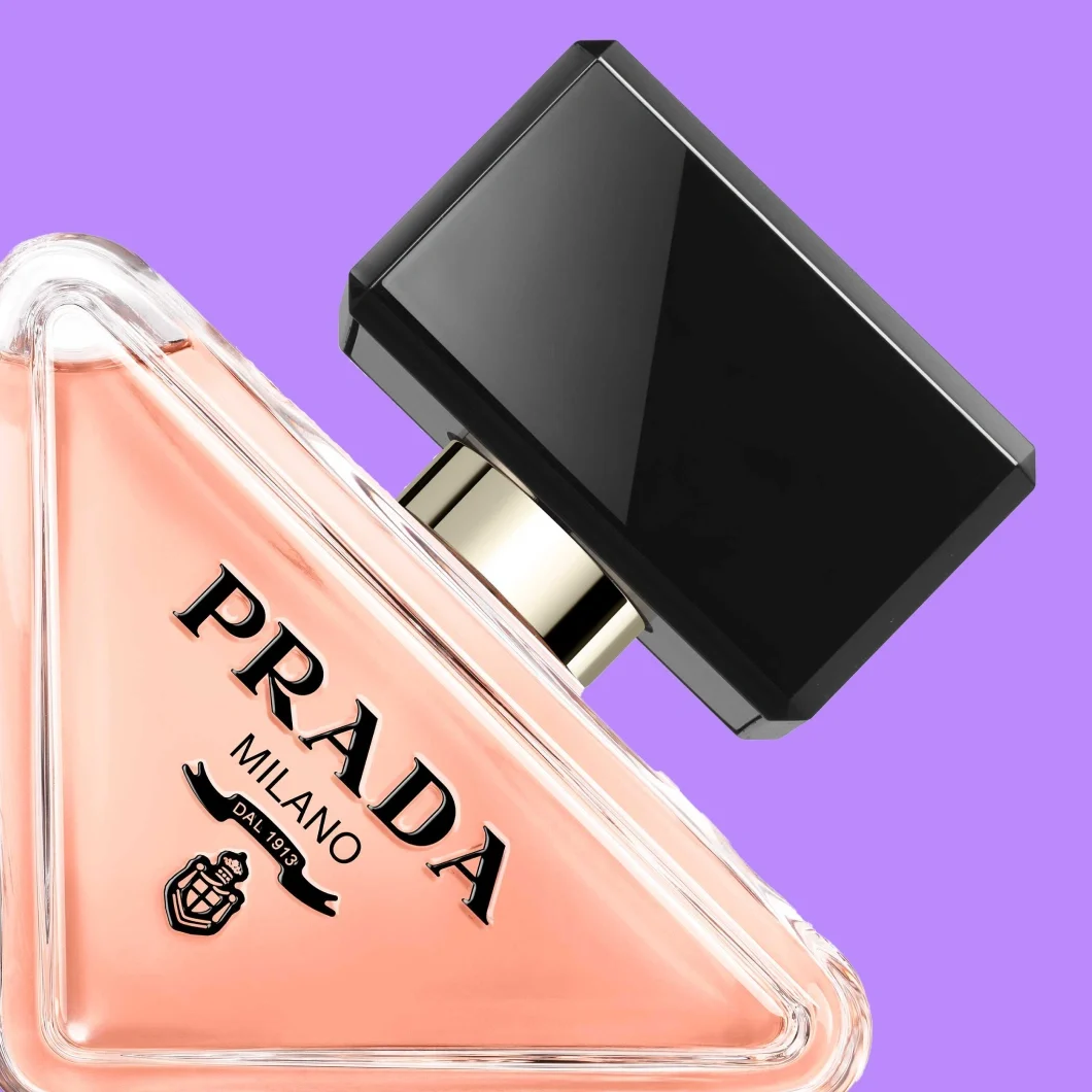 Närbild av Prada Milano-parfym i en triangelformad transparent flaska med ljusrosa innehåll, svart geometrisk kork och synlig text ”PRADA MILANO DAL 1913”, mot lila bakgrund.