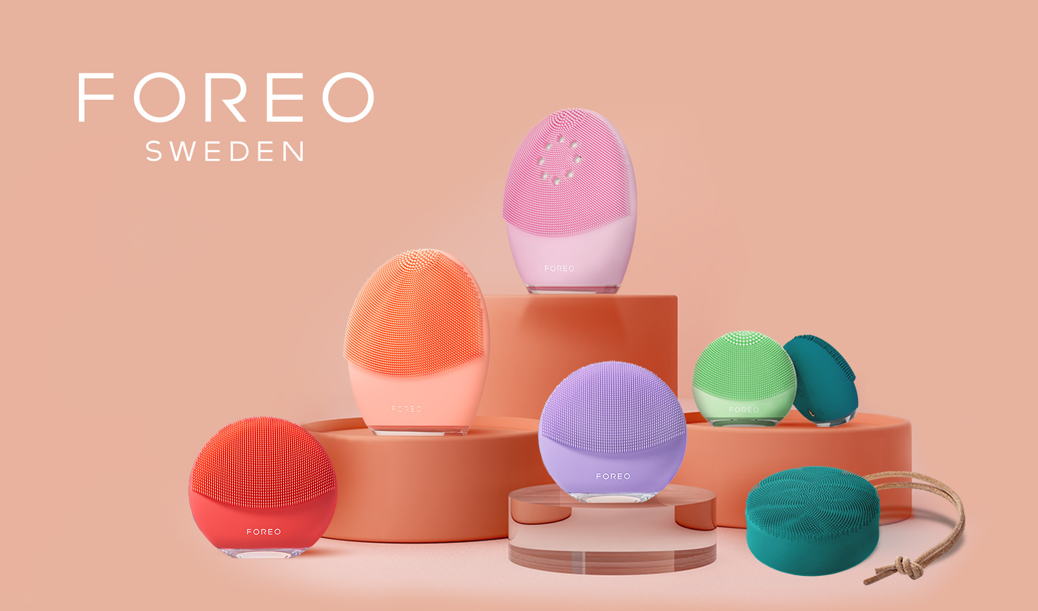 FOREO Sweden -brändin värikkäiden kasvojenpuhdistusaineiden kokoelma on järjestetty kirkkaan oranssille taustalle. Tuotteita on saatavana eri väreissä, kuten punaisena, oranssina, violettina, pinkkinä, vihreänä ja turkoosina. Kuvan vasempaan yläkulmaan on sijoitettu teksti ”FOREO SWEDEN”. Laitteet on sijoitettu tyylikkäästi eri tasoille pyöreille korokkeille.