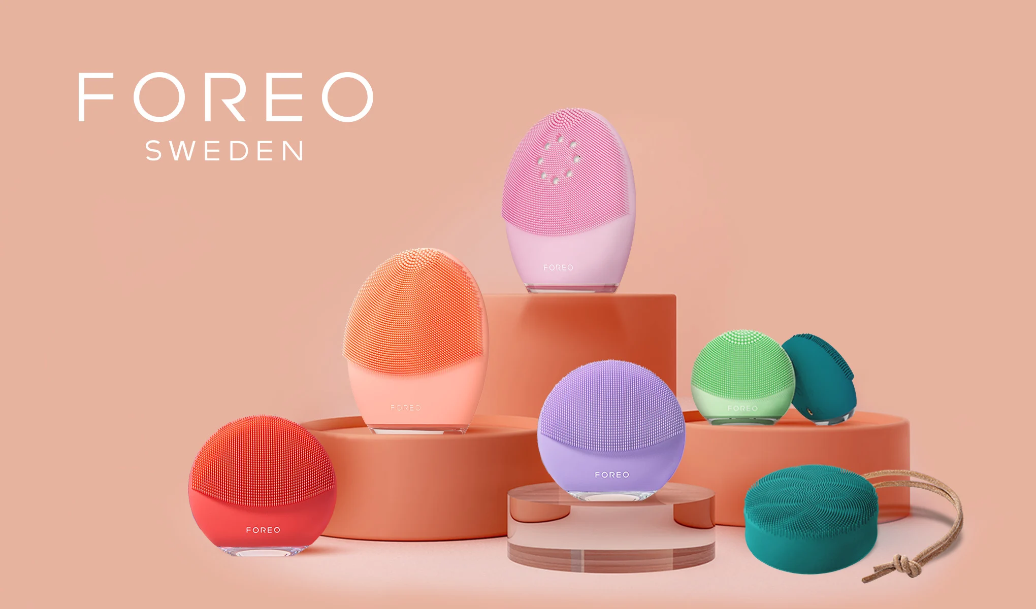 En samling av färgglada ansiktsrengöringsapparater från märket FOREO Sweden är arrangerade på en ljusorange bakgrund. Produkterna finns i olika färger, inklusive röd, orange, lila, rosa, grön och turkos. Texten "FOREO SWEDEN" är placerad i övre vänstra hörnet av bilden. Apparaterna är stilfullt uppställda på olika nivåer av runda podier.