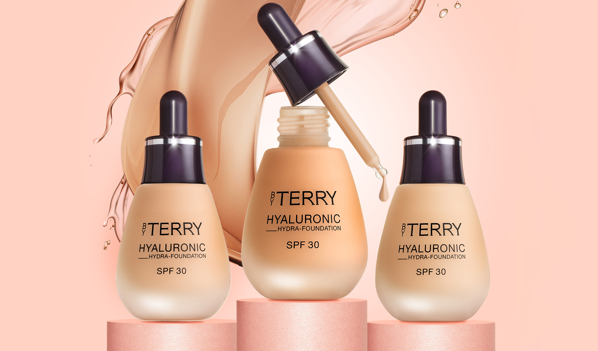 Kuva kolmesta By Terry Hyaluronic Hydra-Foundation SPF 30 -pullosta, jotka on sijoitettu vaaleanpunaisiin sylintereihin. Yhdessä pullossa on pipetti, josta tippuu meikkivoidetta. Tausta on vaaleanpunainen, ja siinä on roiskevaikutelma meikkivoiteesta.