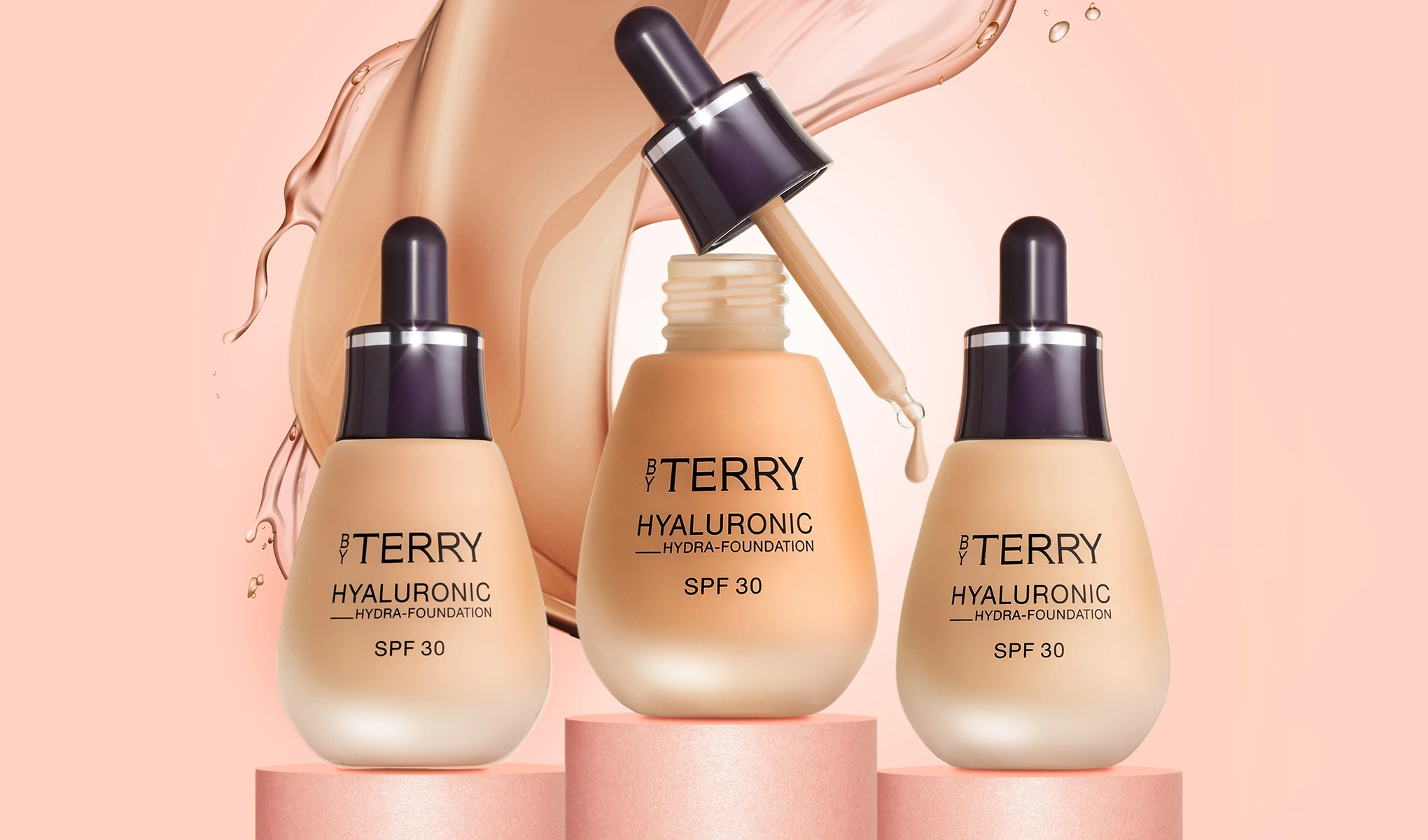 Kuva kolmesta By Terry Hyaluronic Hydra-Foundation SPF 30 -pullosta, jotka on sijoitettu vaaleanpunaisiin sylintereihin. Yhdessä pullossa on pipetti, josta tippuu meikkivoidetta. Tausta on vaaleanpunainen, ja siinä on roiskevaikutelma meikkivoiteesta.