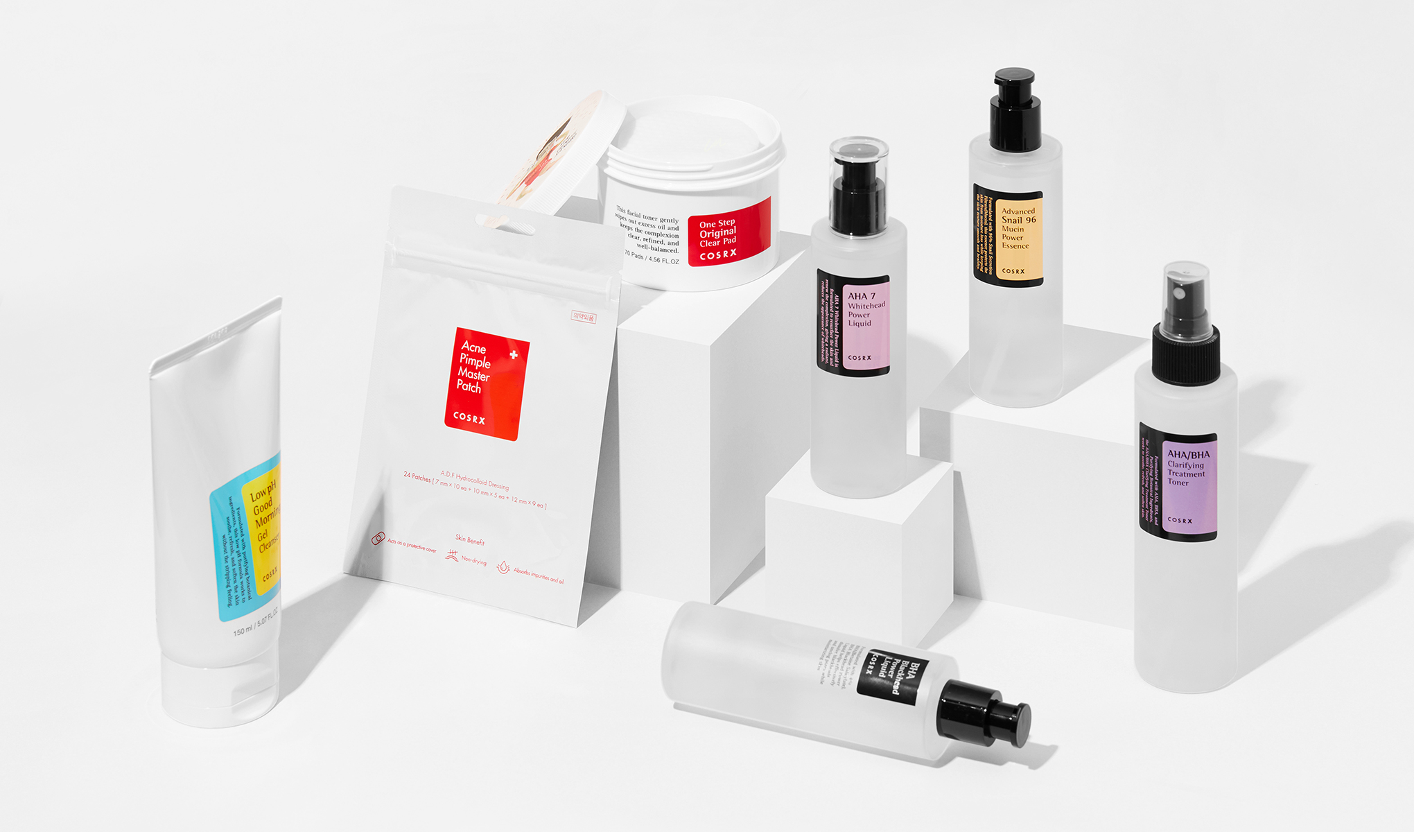 Flere hudpleieprodukter fra merket COSRX. Produktene er plassert på hvite kuber mot en hvit bakgrunn.