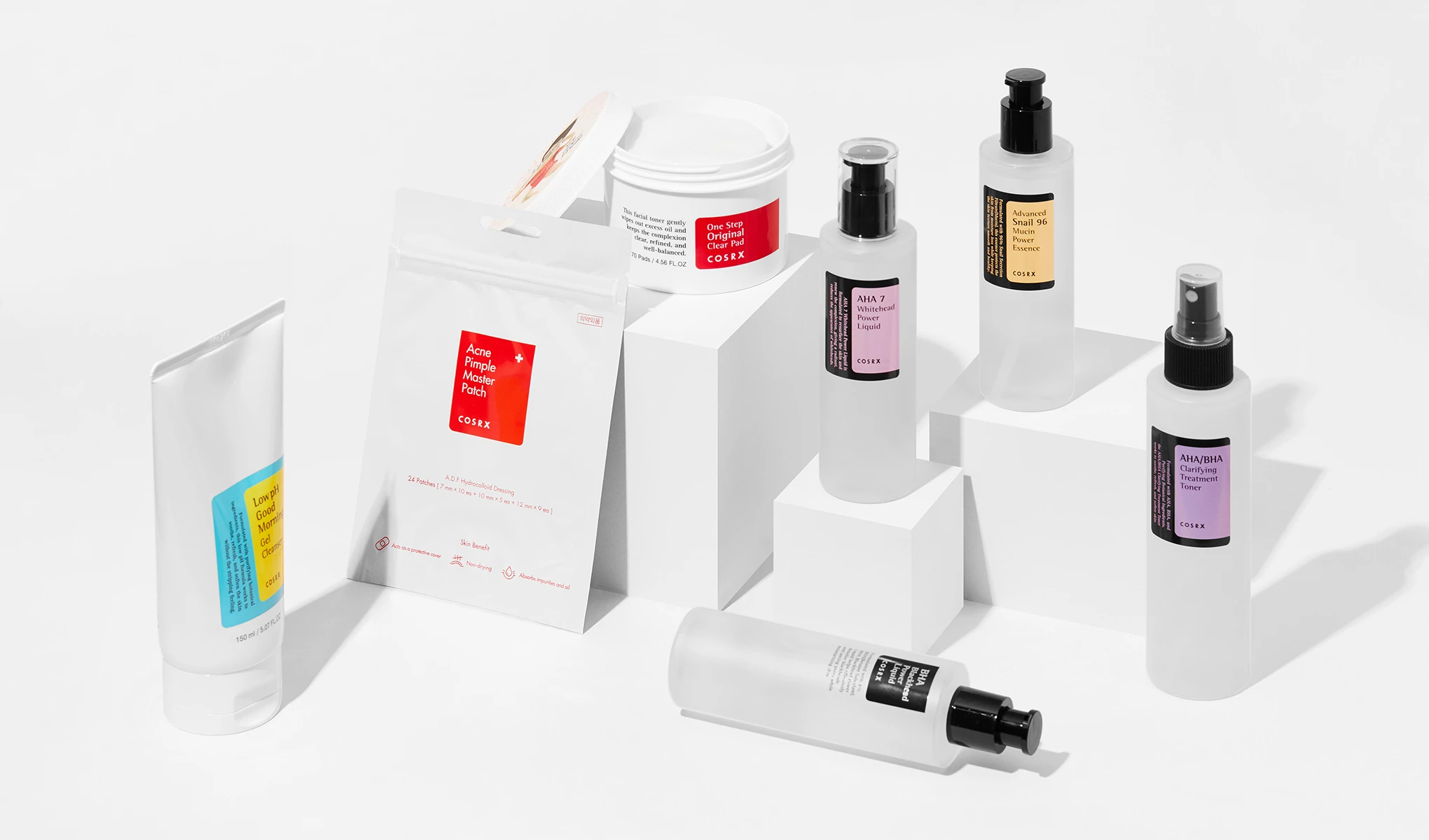 Flere hudpleieprodukter fra merket COSRX. Produktene er plassert på hvite kuber mot en hvit bakgrunn.