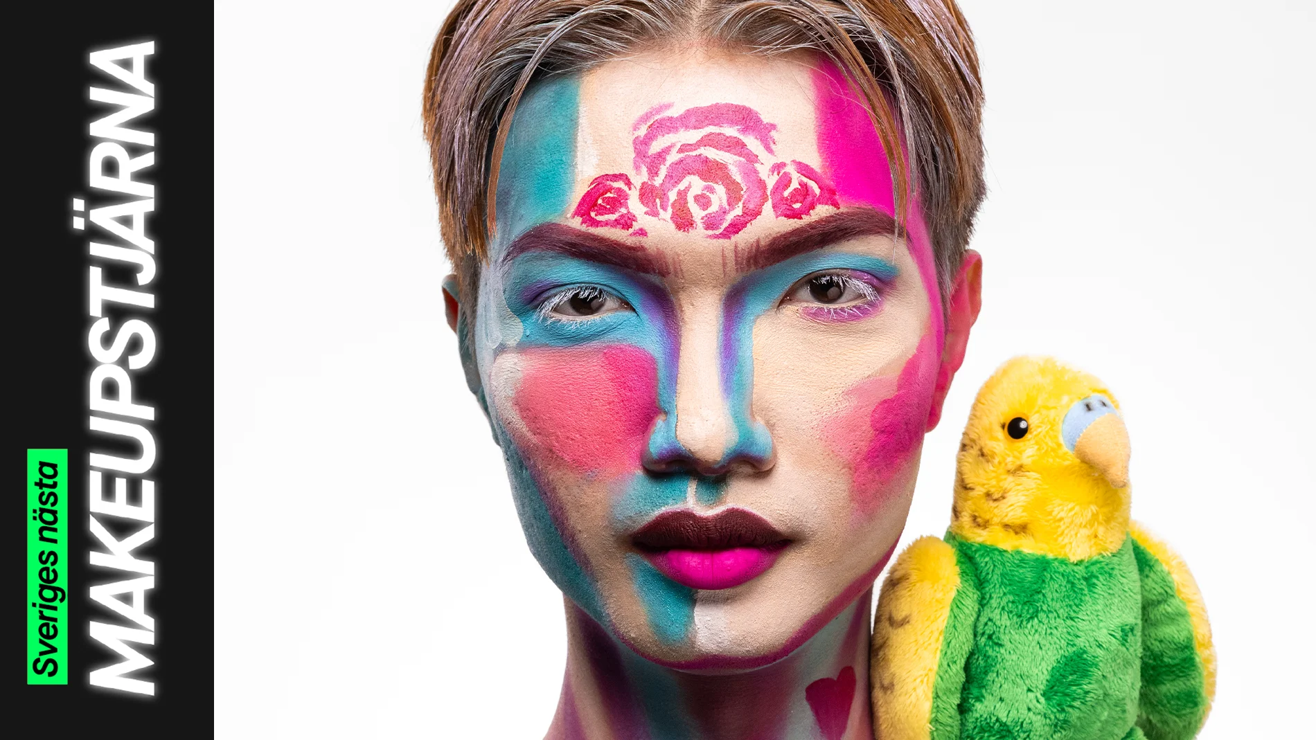 En person med färgstark ansiktsmålning i blå, rosa och lila blockfärger tillsammans med målade rosor i pannan och neonrosa läppar, poserar bredvid en grön och gul gosedjursfågel.