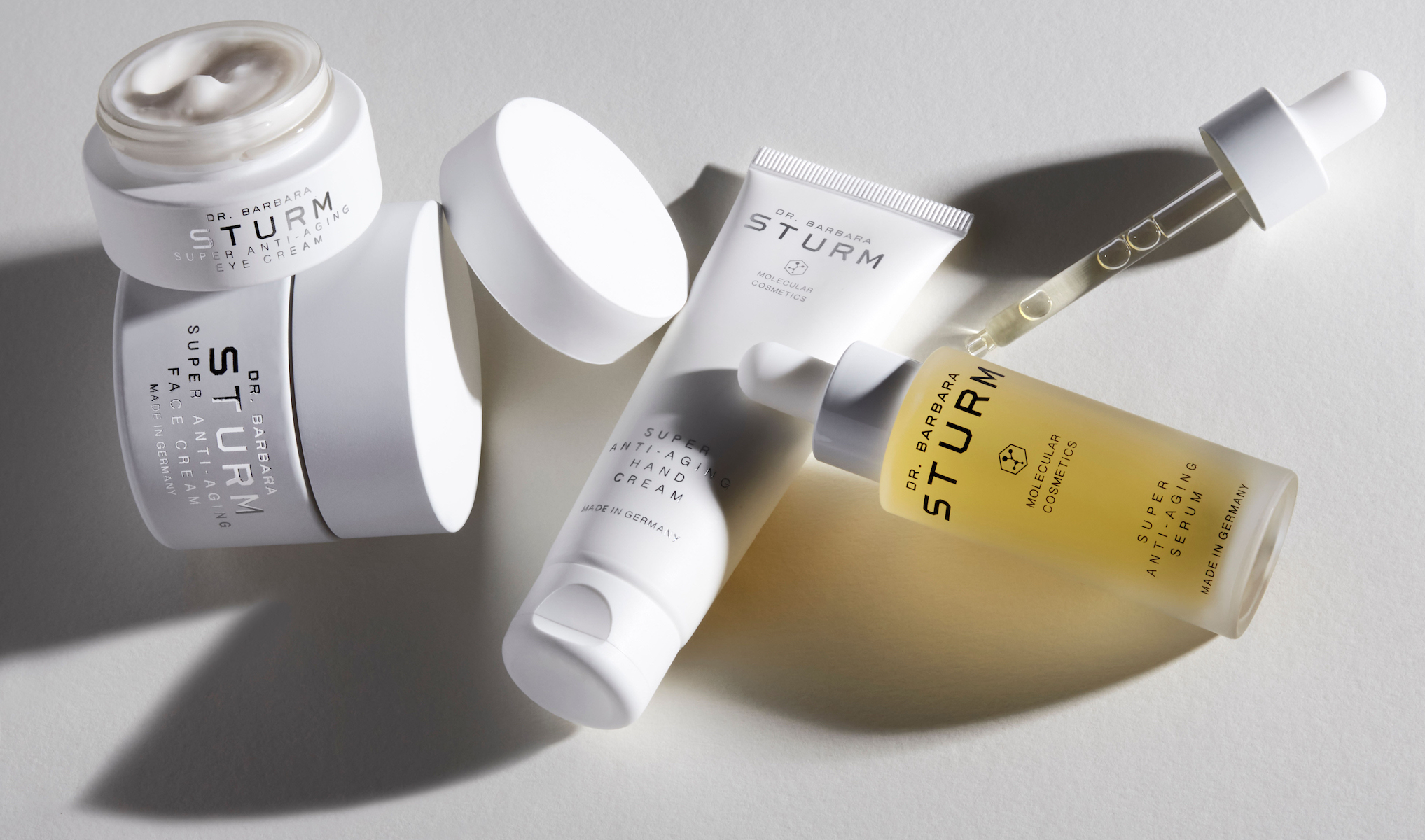 Fyra hudvårdsprodukter från Dr. Barbara Sturm, en burk, en tub, en flaska med en pipett. Produkterna är placerade på en ljus yta och har en minimalistisk design med vita och genomskinliga förpackningar.