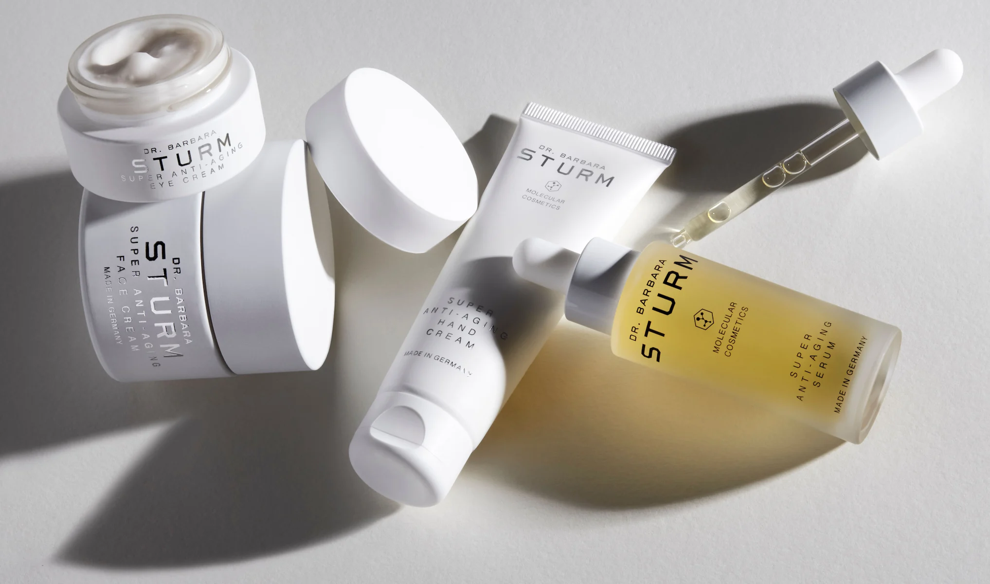 Fyra hudvårdsprodukter från Dr. Barbara Sturm, en burk, en tub, en flaska med en pipett. Produkterna är placerade på en ljus yta och har en minimalistisk design med vita och genomskinliga förpackningar.