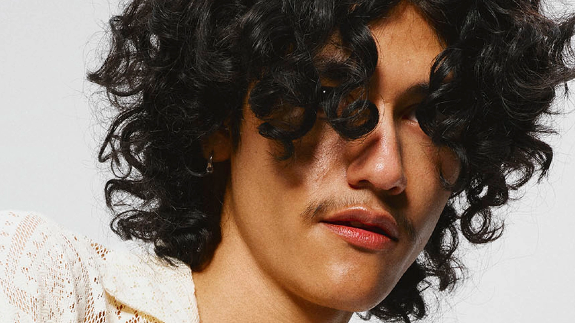 Nærbilde av en person med mørkt, krøllete hår som faller over pannen og rundt ansiktet. Personen har på seg en lys, strukturert skjorte mot en nøytral bakgrunn.