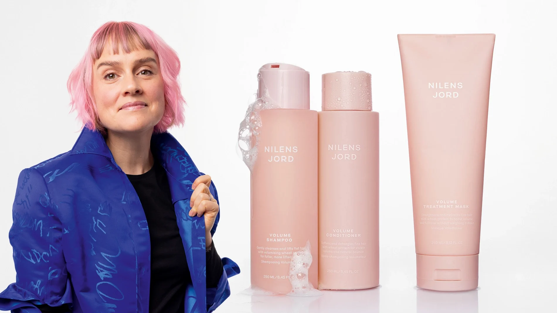 Kvinne med rosa hår i blå jakke ved siden av tre rosa Nilens Jord Volume hårprodukter: shampoo, balsam og hårkur.