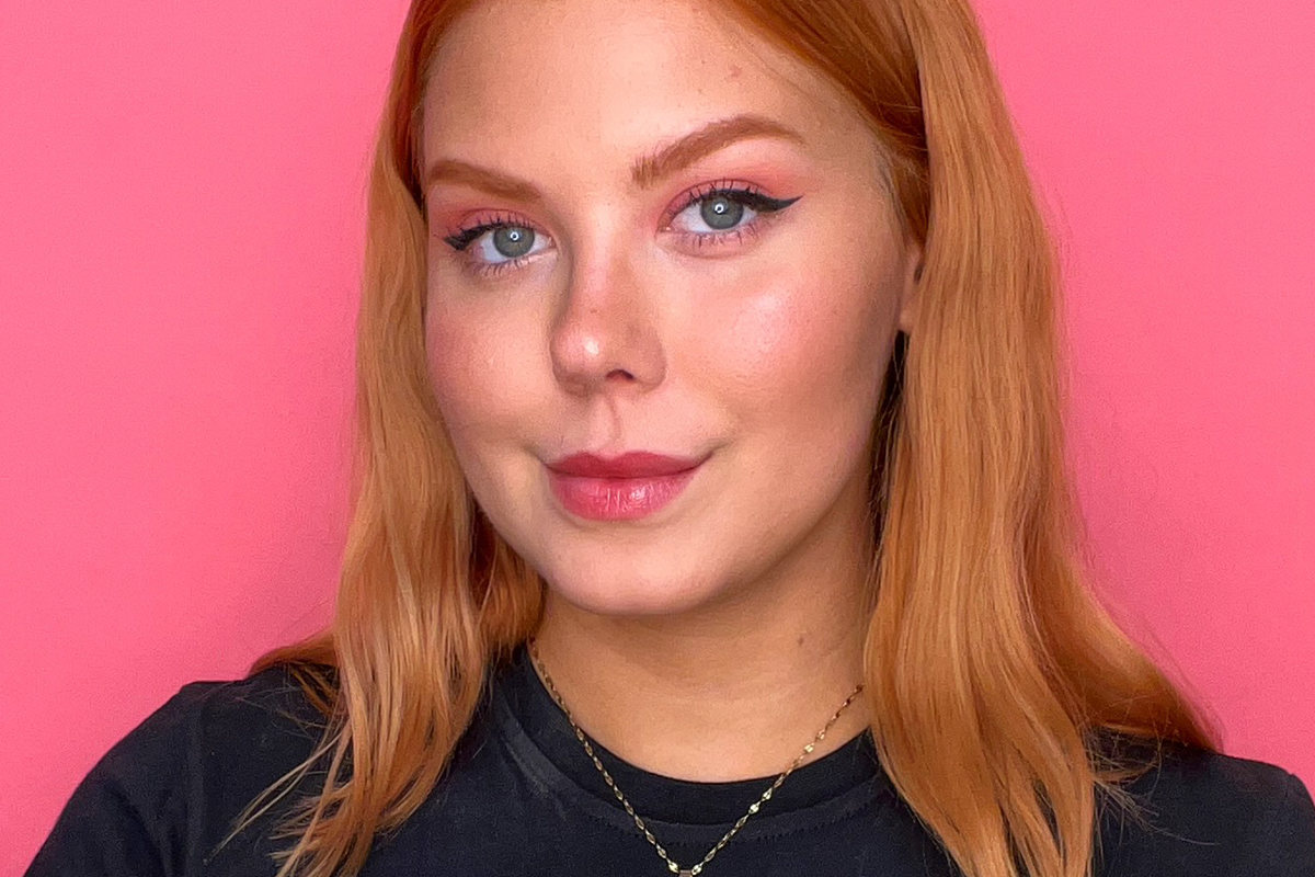  Lähikuva hymyilevästä naisesta, jolla on punertavanvaaleat hiukset ja valmis, luonnollinen meikki, johon kuuluu eyeliner, poskipuna ja huuliväri.