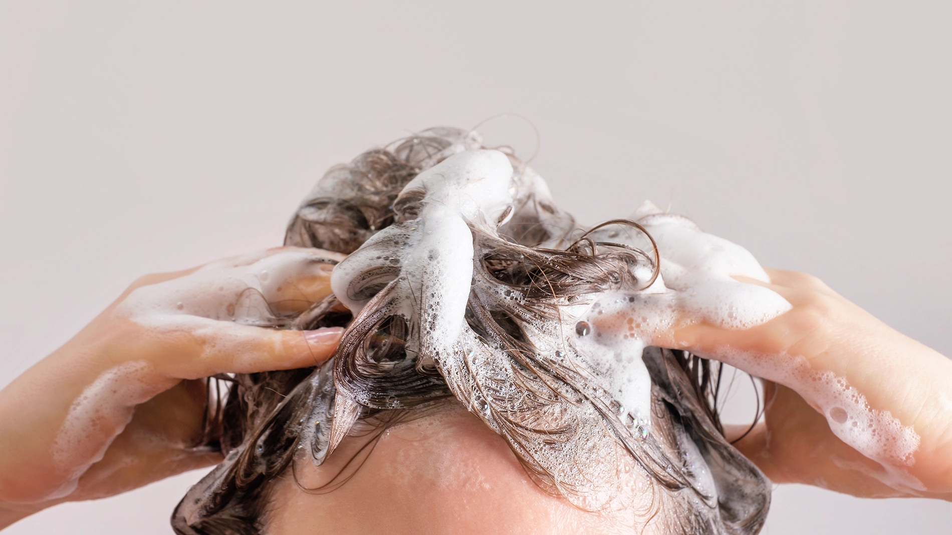 Närbild på en person som tvättar håret med schampo, händer masserar hårbotten och blött hår täckt av vitt lödder mot ljus bakgrund.