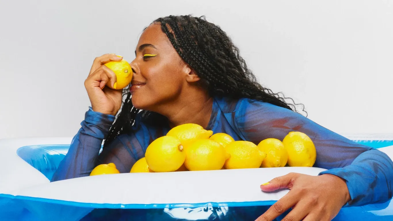 Kvinna med ljusgul eyeliner som har massor av citron framför sig. Kvinnan luktar på en citron och ler.
