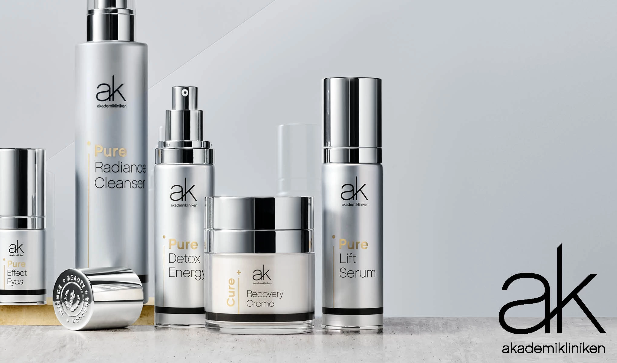 Akademiklinikenin ihonhoitotuotteita, kuten Pure Radiance Cleanser, Pure Detox Energy, Pure Lift Serum, Pure Effect Eyes ja Cure Recovery Creme. Tuotteet on sijoitettu vaalealle alustalle, jonka oikeassa alakulmassa on Akademiklinikenin logo.