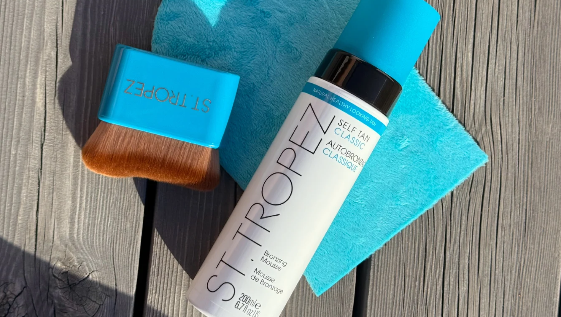 St.Tropez Self Tan Classic Bronzing Mousse, 200 ml, med texten ”Natural Healthy Looking Tan”, tillsammans med en turkos St.Tropez appliceringsborste och en turkos handduk på ett träunderlag.