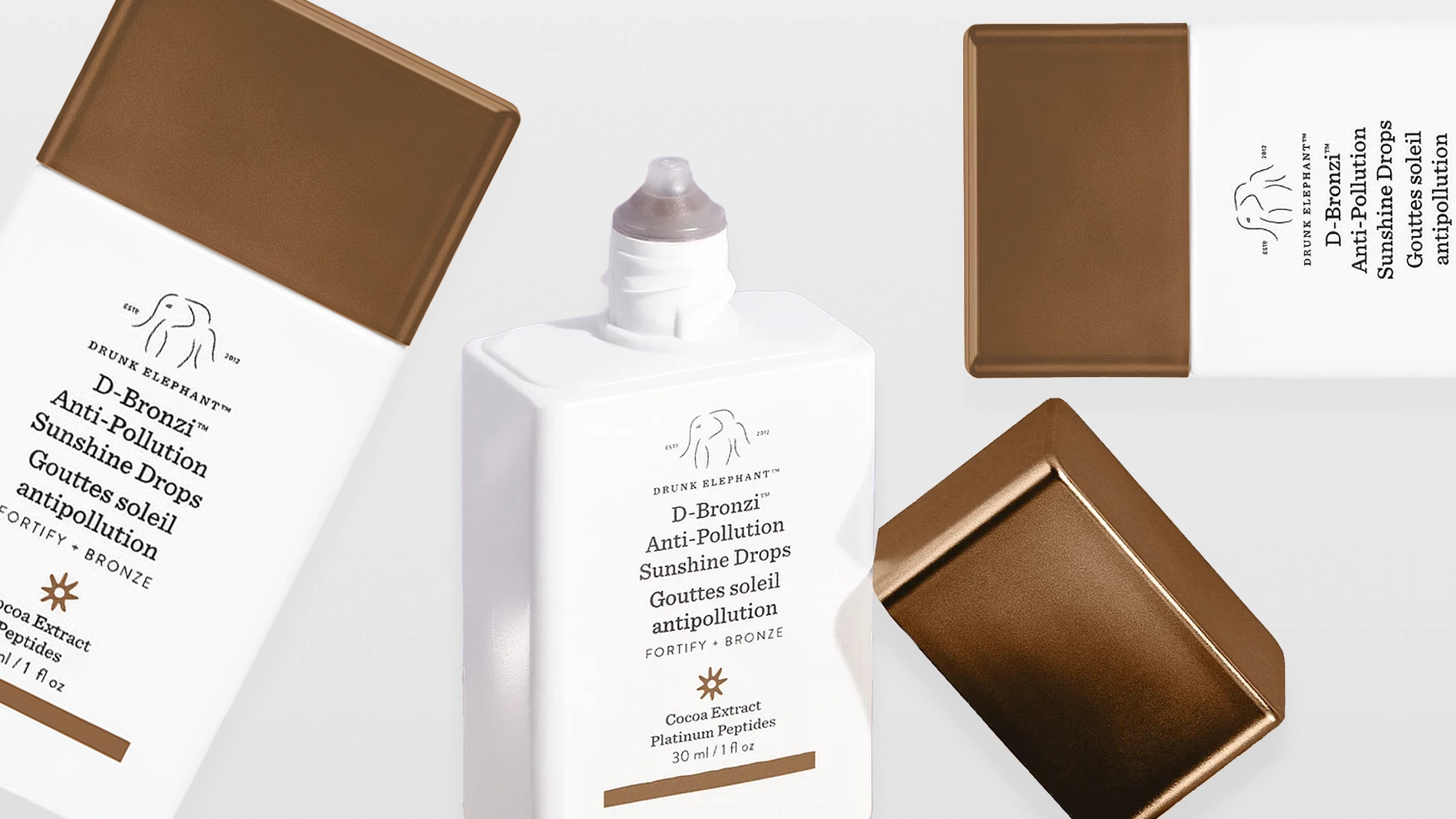 Tuotekuva, jossa useita Drunk Elephant D-Bronzi Anti-Pollution Sunshine Drops -pulloja ja korkkeja. Valkoisia, neliskulmaisia pulloja pronssinvärisillä korkeilla eri asennoissa vaaleaa taustaa vasten.