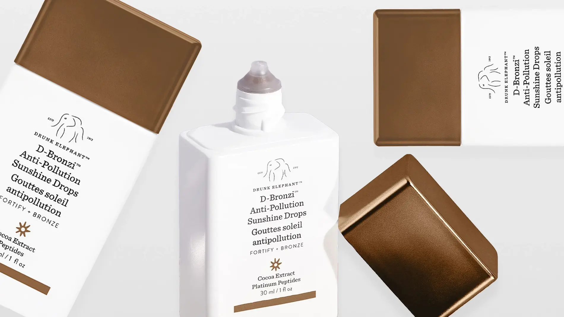 Tuotekuva, jossa useita Drunk Elephant D-Bronzi Anti-Pollution Sunshine Drops -pulloja ja korkkeja. Valkoisia, neliskulmaisia pulloja pronssinvärisillä korkeilla eri asennoissa vaaleaa taustaa vasten.