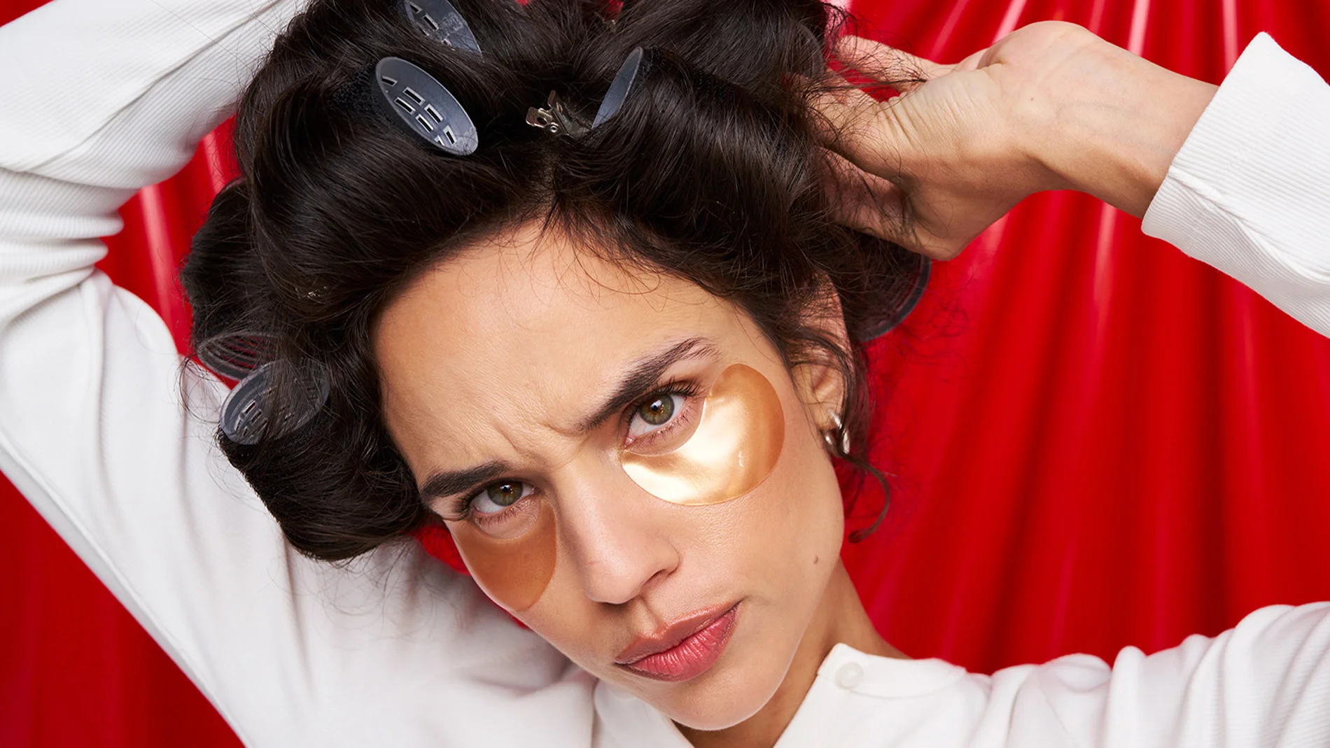 Kvinne med mørkt hår i hårruller og gullfargede øyemasker under øynene står foran en rød bakgrunn. Hun holder hendene i håret og ser konsentrert mot kameraet.