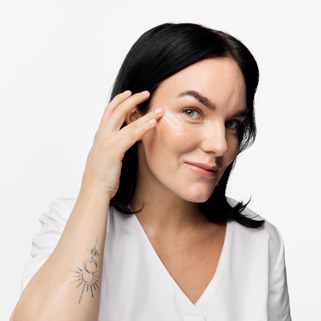 Person med svart hår påfører øyekrem under øyet mot lys grå bakgrunn og har på seg en hvit topp. Synlig tatovering på underarmen med sol- og månemotiv.