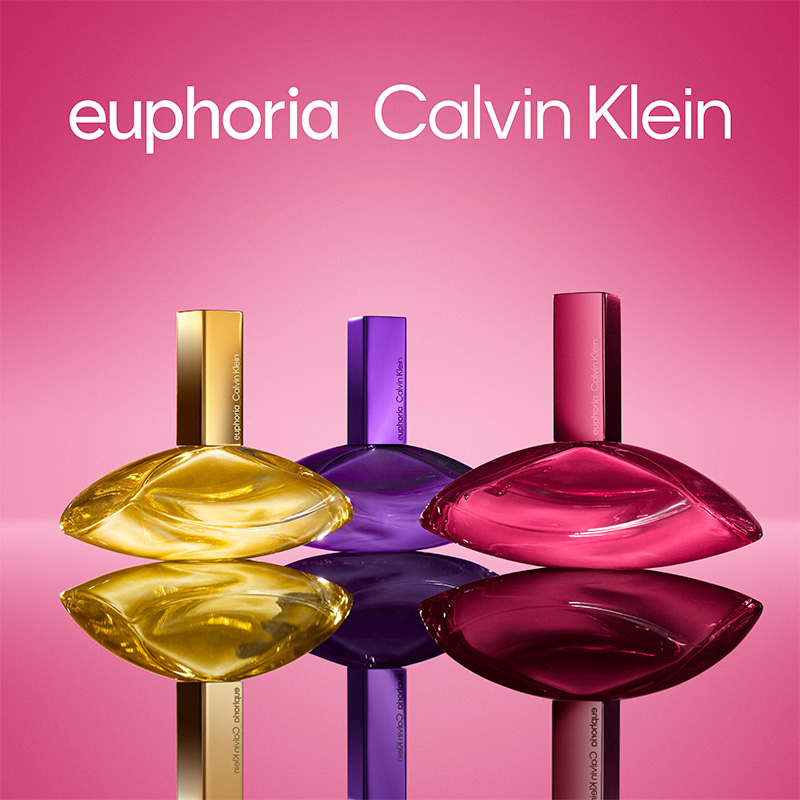 Calvin Klein Euphoria. Tre parfymflaskor i guld, lila och rosa mot rosa bakgrund. Flaskorna speglas i underlaget.
