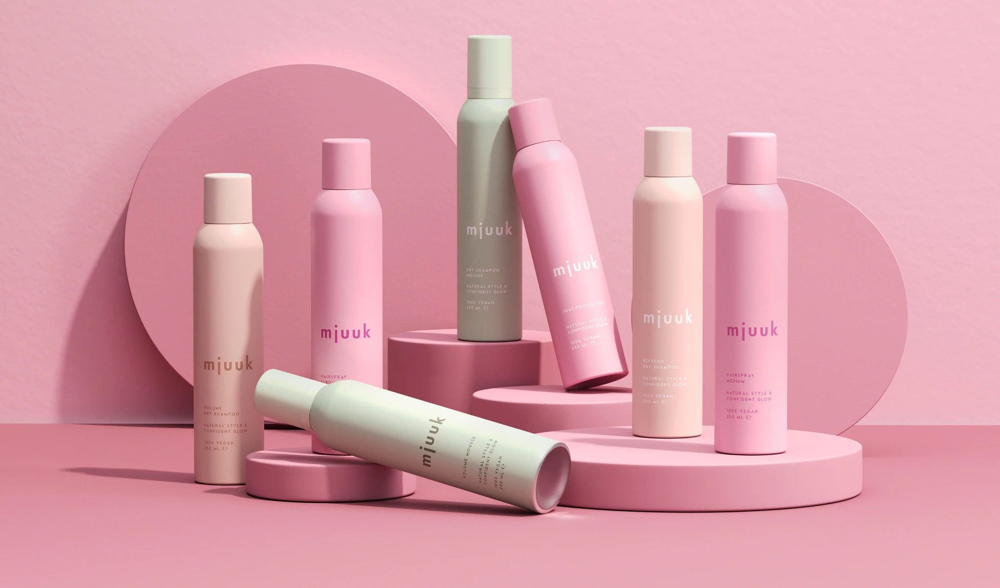 Flere rosa og pastellfargede hårprodukter er stilfullt plassert på en rosa bakgrunn. Produktene er av merket "mjuuk" og inkluderer blant annet tørrsjampo og hårspray.